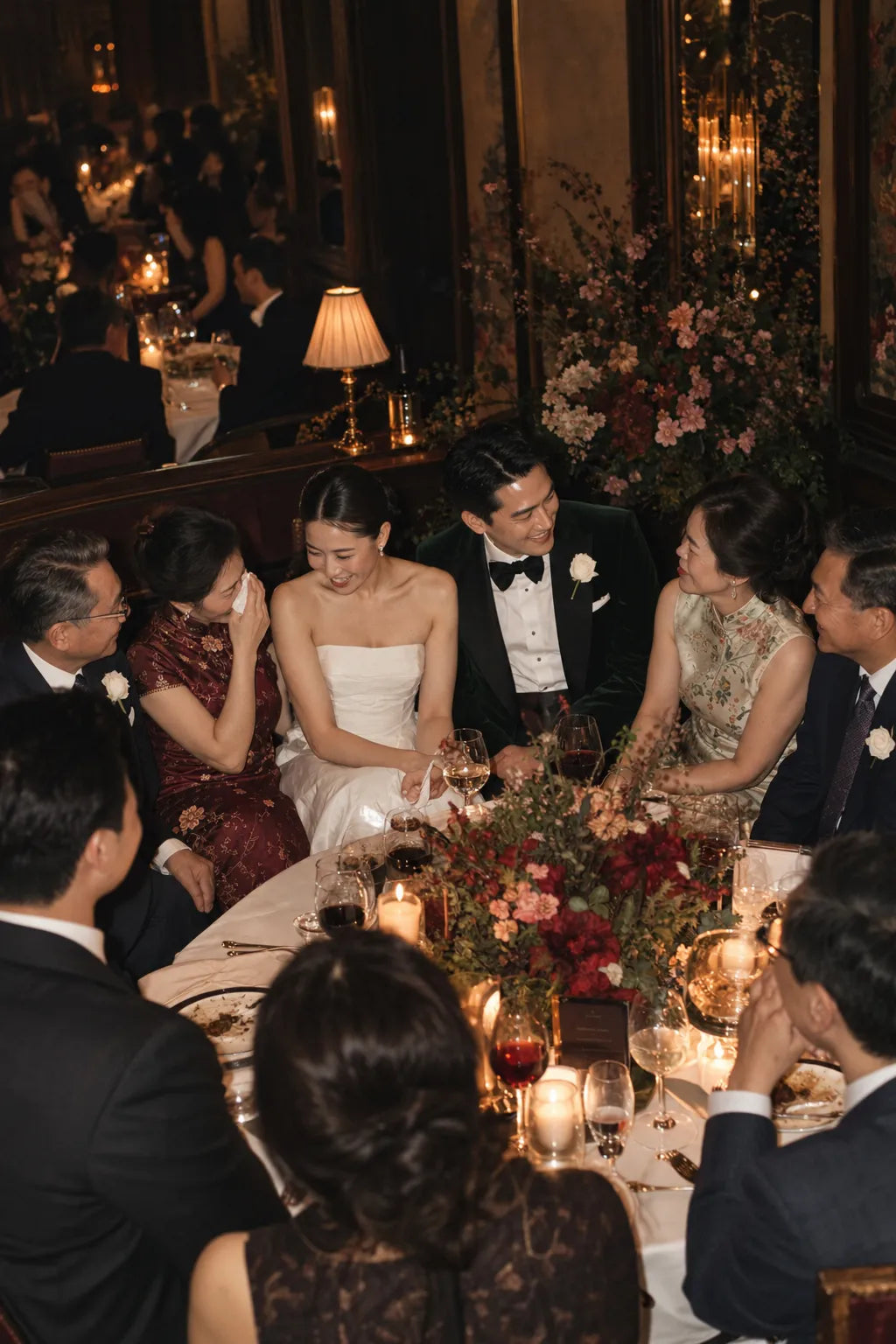 A candid wedding reception scene showing a bride in a white strapless gown and a groom in a black tuxedo sitting at a candlelit dinner table surrounded by guests.