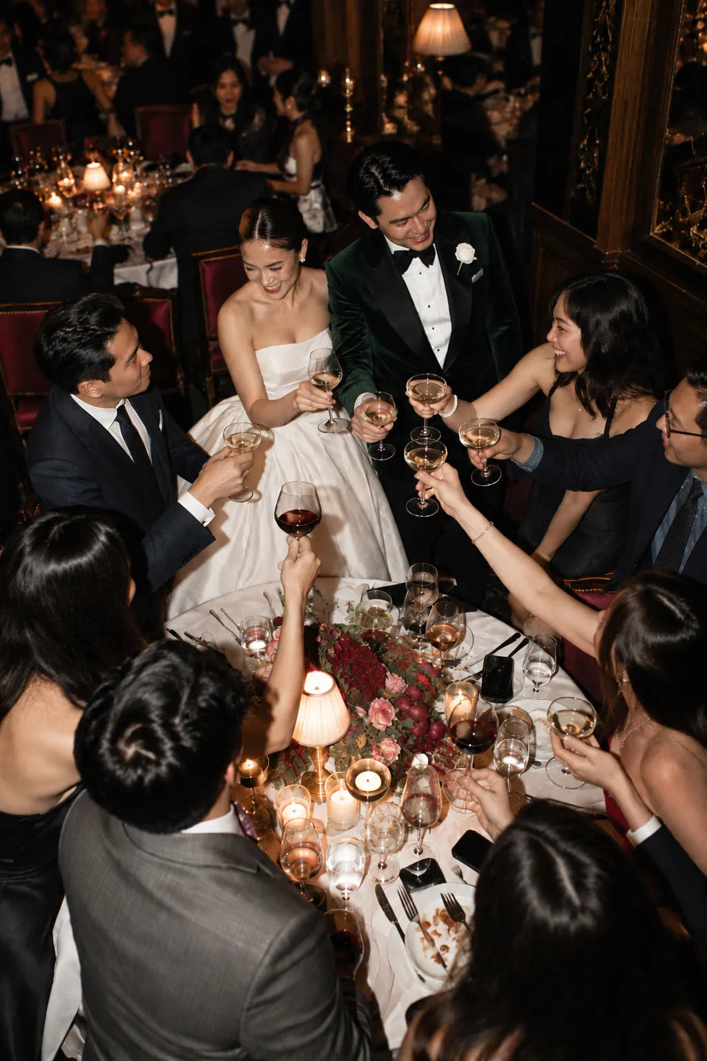 A bride in a white strapless gown and a groom in a dark green tuxedo with a bow tie are seated at a wedding reception table, surrounded by guests raising glasses of wine during a toast.