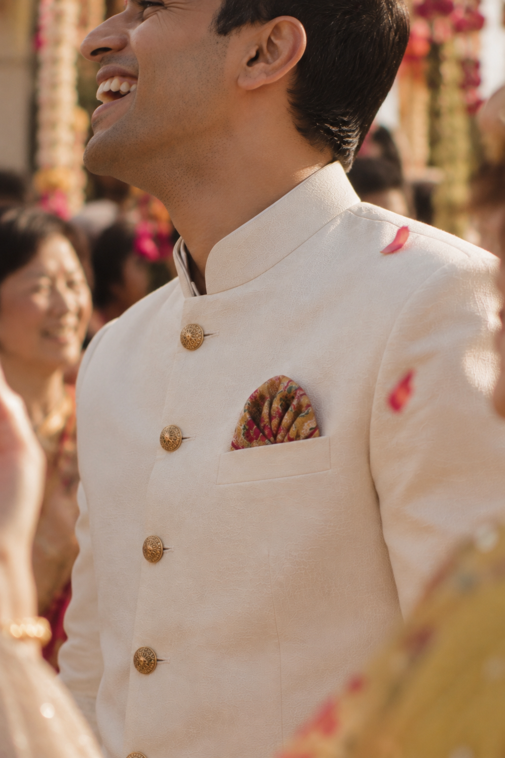 A man in a cream-colored traditional Indian sherwani jacket with gold buttons and a patterned pocket square, smiling in a festive outdoor setting.