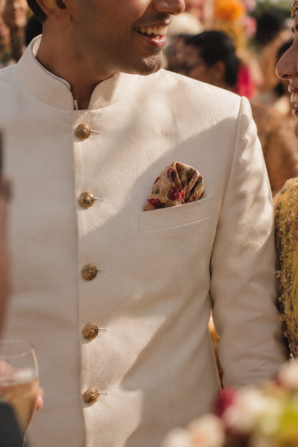 A close-up of a man wearing a cream-colored Indian sherwani jacket with gold buttons and a patterned pocket square, set against a warm, festive background.