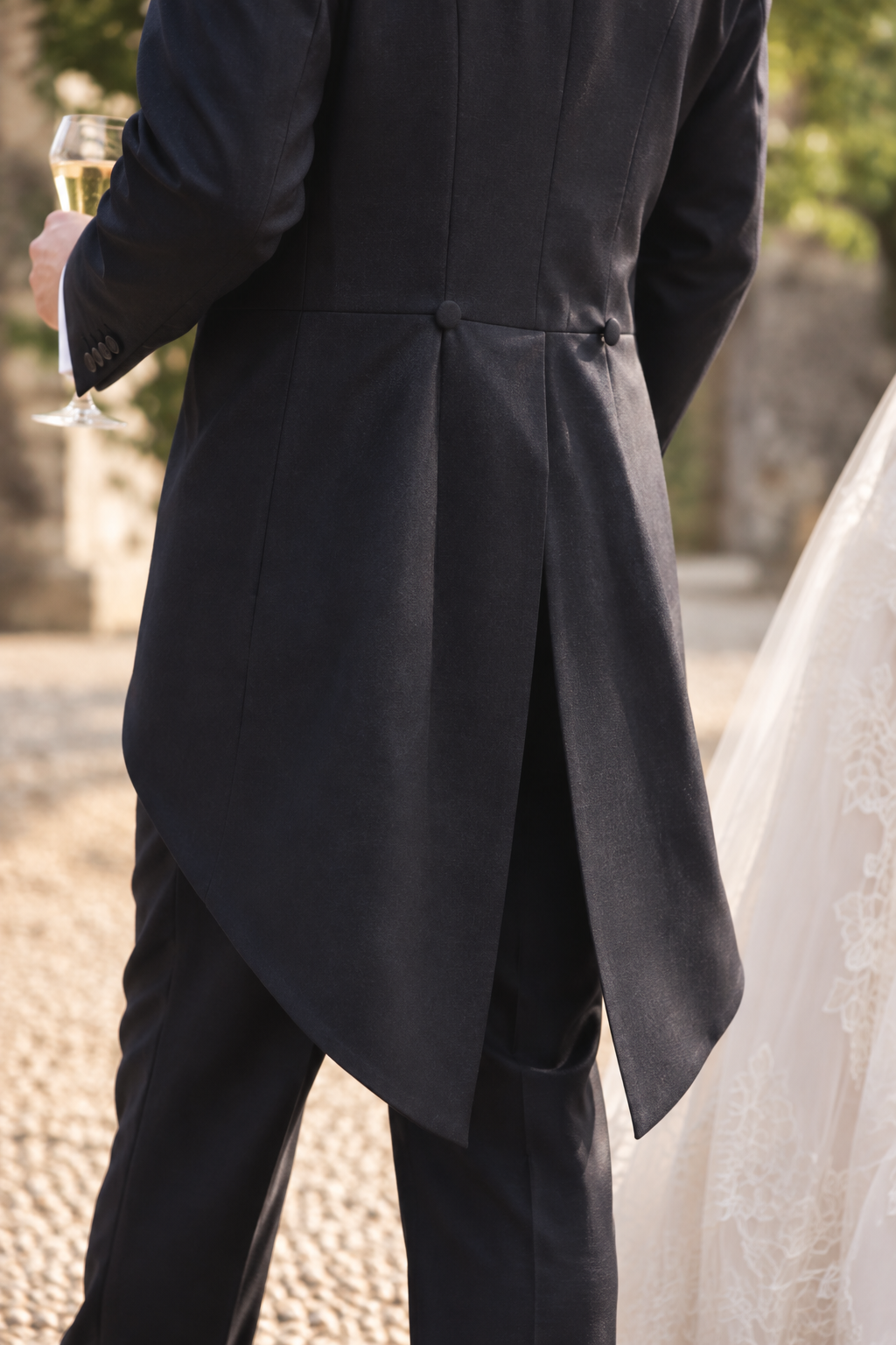 Rear view of a man in a dark formal tailcoat and trousers, holding a glass of champagne, with part of a bride's white lace dress visible at the right edge in an outdoor setting.