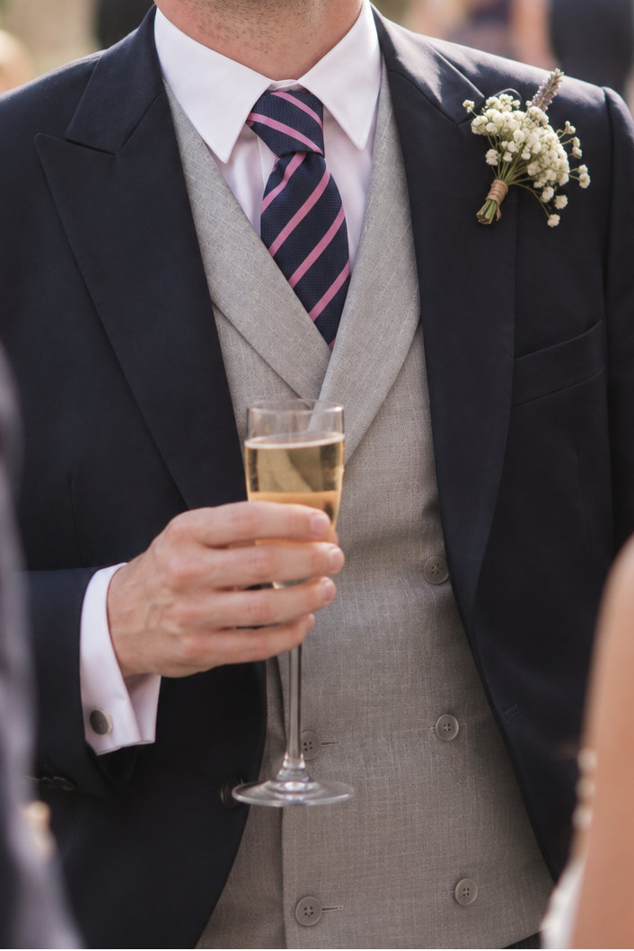 A man in a dark navy suit, grey waistcoat, and striped pink tie laughs while holding a glass of champagne at an outdoor wedding reception.