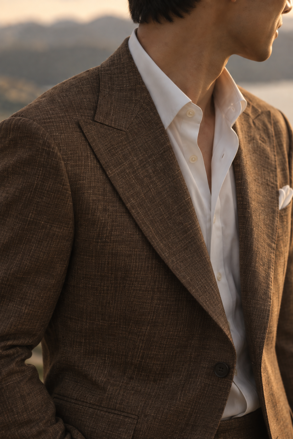 A close-up of a man wearing a brown textured blazer over a white dress shirt, with a blurred outdoor mountain background.