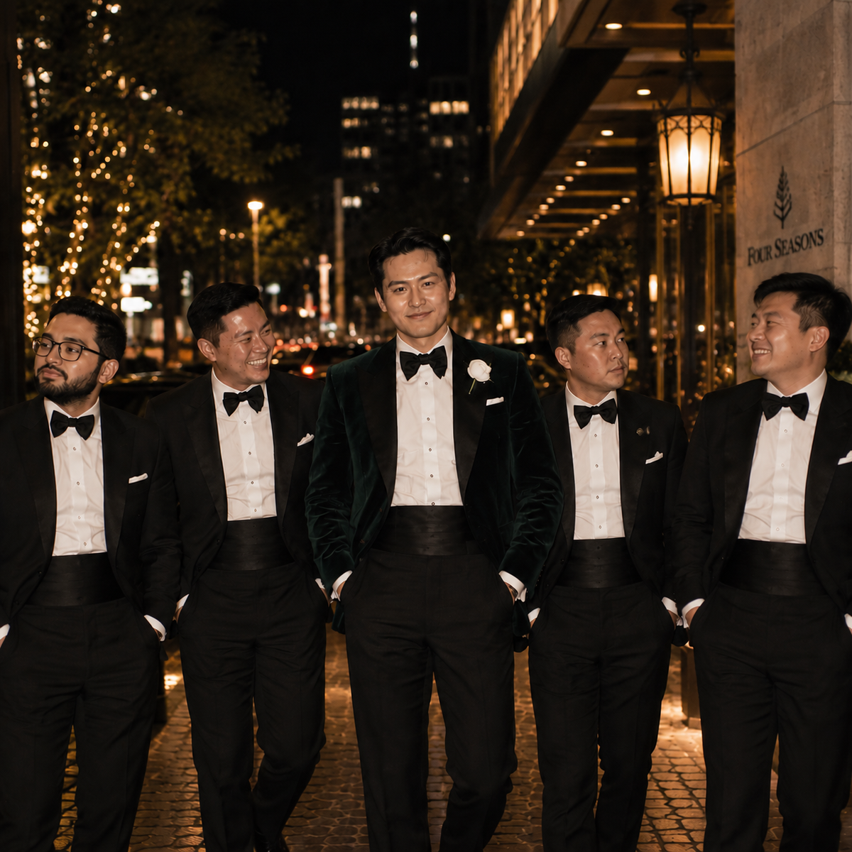 Five men in matching black tuxedos with white shirts and black bow ties walk together at night on a cobblestone street. The central man wears a black velvet tuxedo jacket and a white boutonniere.