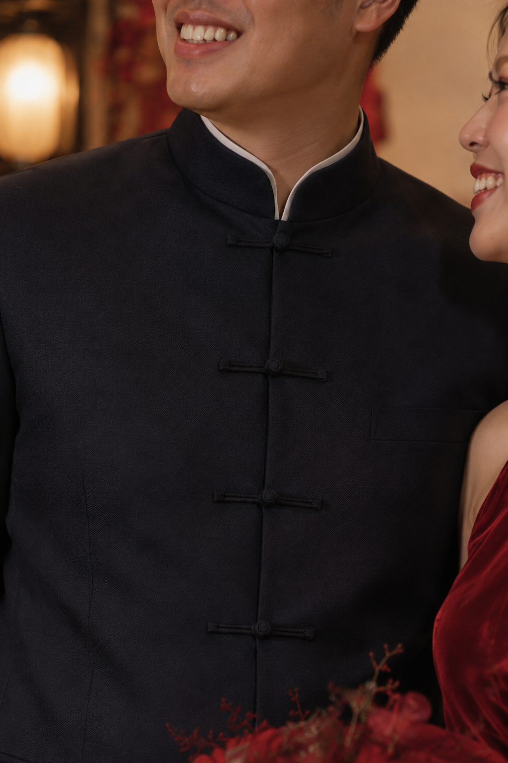 A close-up lifestyle shot of a man wearing a dark navy traditional Chinese mandarin jacket with black buttons and a white inner collar, standing next to a woman in a red dress.