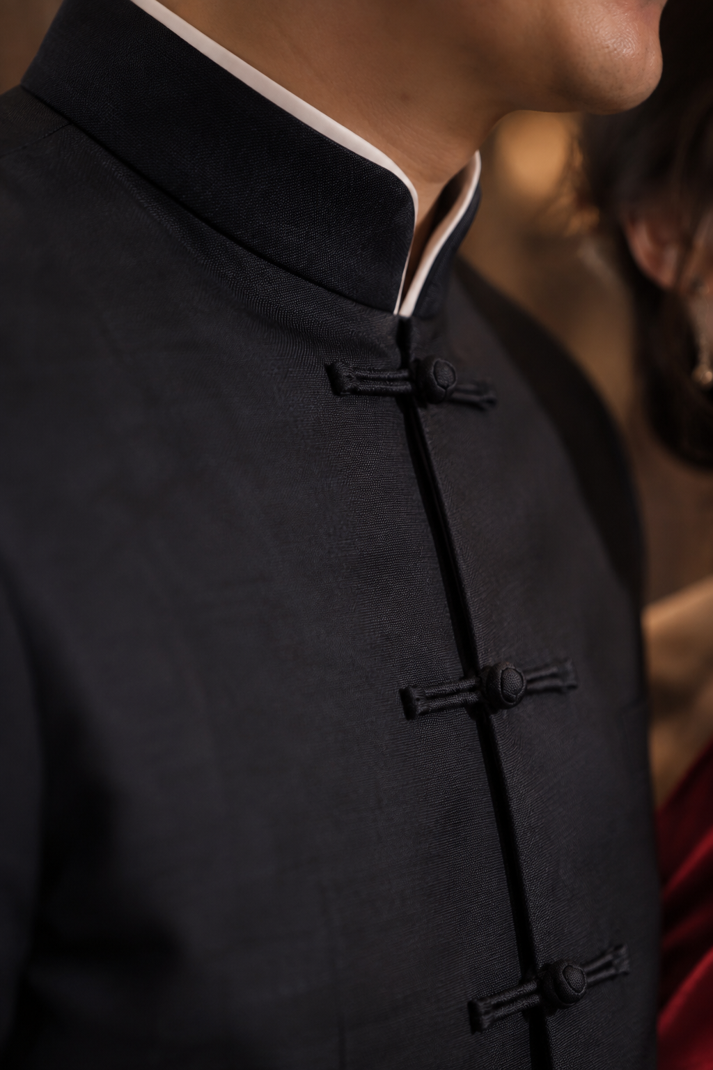 Close-up of a person wearing a dark navy traditional Chinese-style jacket with a mandarin collar and black button closures, against a softly blurred indoor background.
