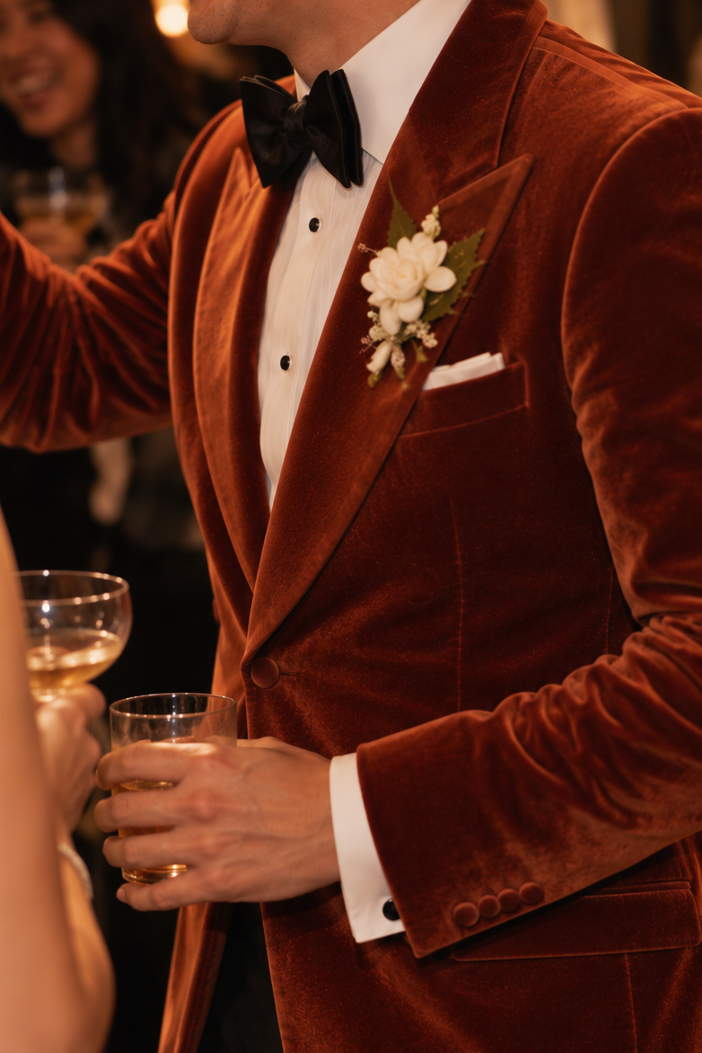 A man wearing a rich rust-brown velvet tuxedo jacket with a white shirt, black bow tie, and a white floral boutonniere, captured at a warm, celebratory event.