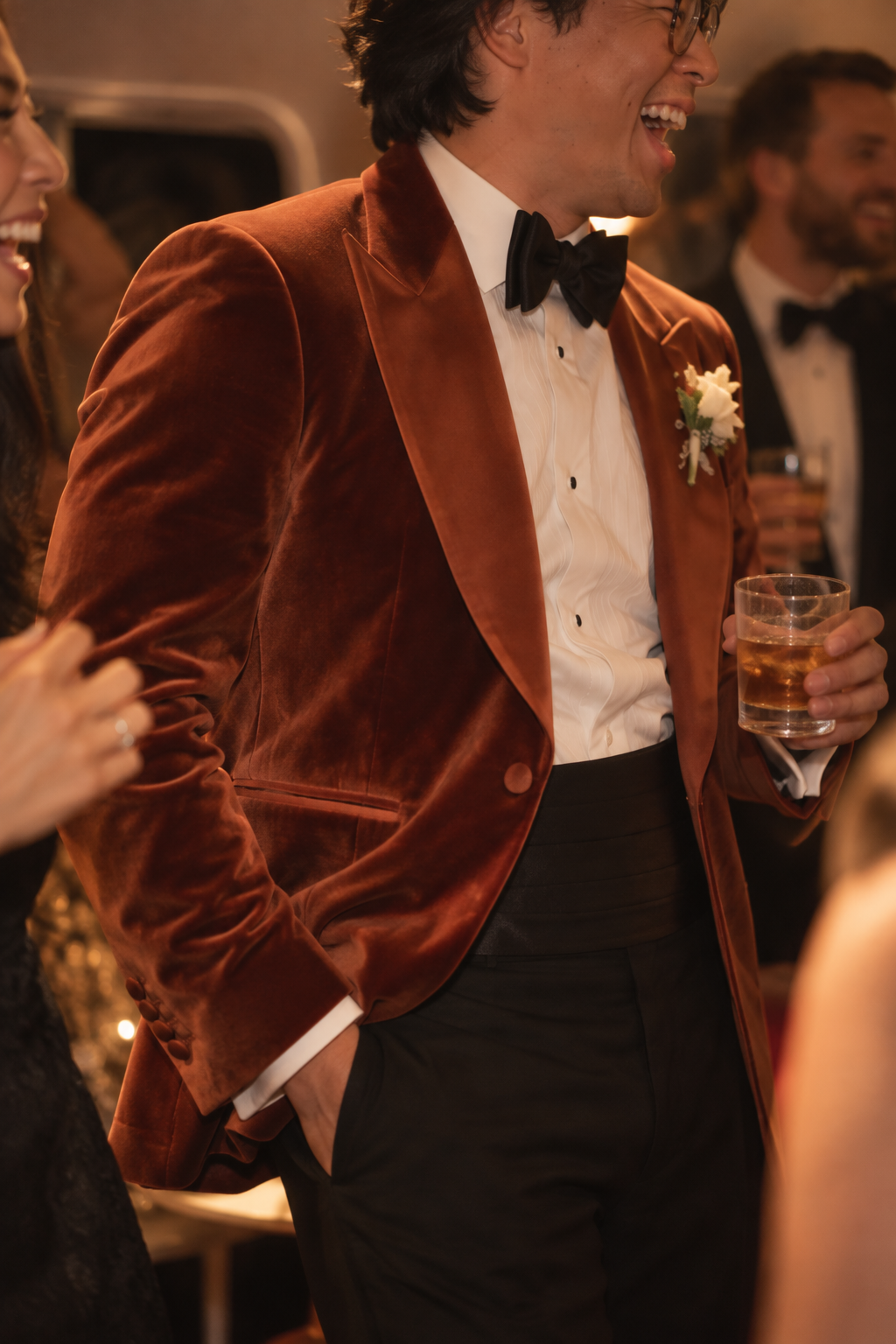 A man at a formal event wearing a rust-brown velvet tuxedo jacket over a white dress shirt and black bow tie, holding a glass of whiskey.